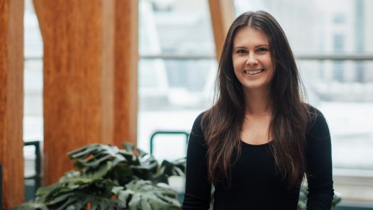 A woman with long brown hair, wearing a black long-sleeve top and a belt, stands in a bright, modern space with large wooden beams and windows in the background. She is smiling at the camera, with greenery visible in the foreground.