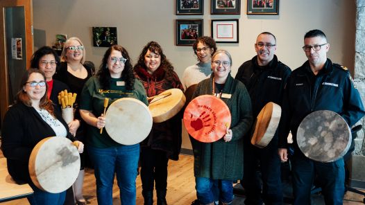 Group of nine people stand in UNBC's First Nations Centre holding hand drums