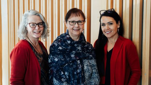 Three women standing in front of a wood-panelled wall. From left to right: Margot Parkes, Lheidli T'enneh Elder and UNBC Chancellor Darlene McIntosh, and Rheanna Robinson. They are smiling and wearing red and dark-coloured clothing.