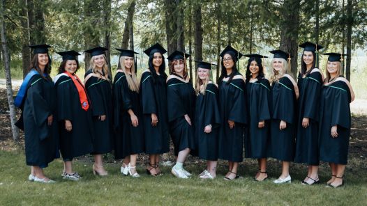 A group of graduates wearing black gowns and mortarboards stand side by side outdoors in front of a backdrop of tall trees. They are smiling and dressed for a graduation celebration, with some wearing hoods or stoles in various colours.