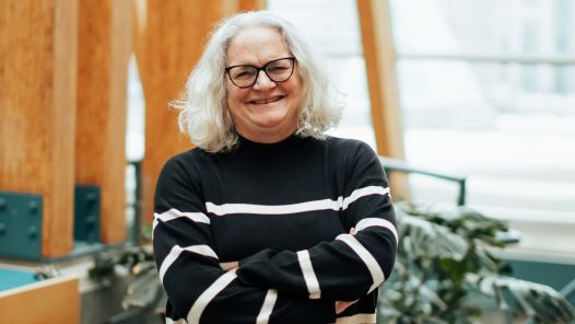 A person with shoulder-length wavy grey hair and black-framed glasses is standing with their arms crossed, smiling. They are wearing a black sweater with white horizontal stripes. The background features a modern indoor space with large wooden beams, glass windows, and green plants.