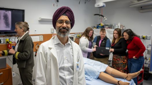 "A man wearing a white lab coat and a maroon turban stands in the foreground of a medical simulation lab, smiling at the camera. Behind him, a group of people is gathered around a hospital bed with a medical mannequin, discussing something on a laptop. A woman in the background is interacting with a screen displaying a chest X-ray."