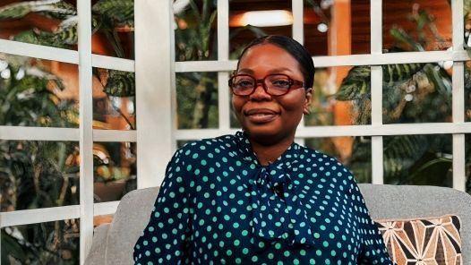 Person wearing glasses and navy polka dot blouse sits on a couch with trellis and plants in background.