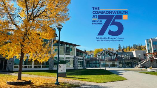 UNBC campus in autumn with golden trees and clear blue skies. The Commonwealth at 75 logo, showing UNBC’s official partnership, is displayed on the right side of the image. The logo states that the publication is produced by St James’s House and the History of Parliament.