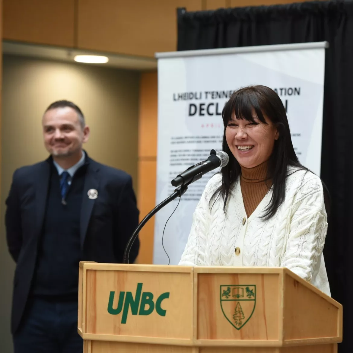 A woman smiles while speaking at a podium with the UNBC logo, during an event indoors. A man in formal attire stands in the background, also smiling. A banner with the title "Lheidli T’enneh First Nation Declaration" is partially visible behind them.