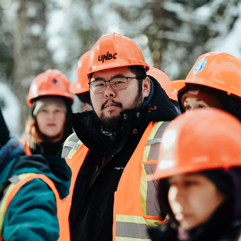 A group of individuals wearing orange safety vests and UNBC-branded hard hats stand outdoors in a snowy environment. A person in the foreground looks directly at the camera, while others in the group appear engaged in conversation or looking around. Trees and a wintery background are visible.