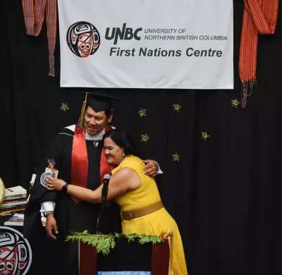 Two people hug at a podium, sign in the background says First Nations Centre. 
