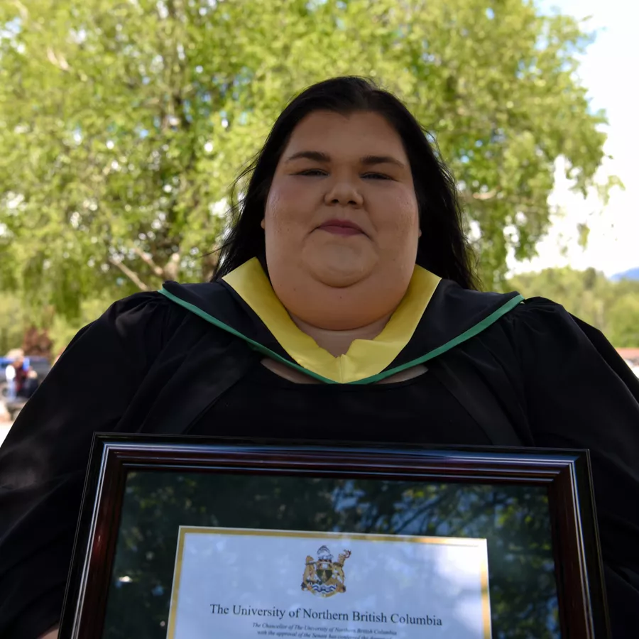 Student wearing black academic robe with yellow striped hood poses holding framed parchment.