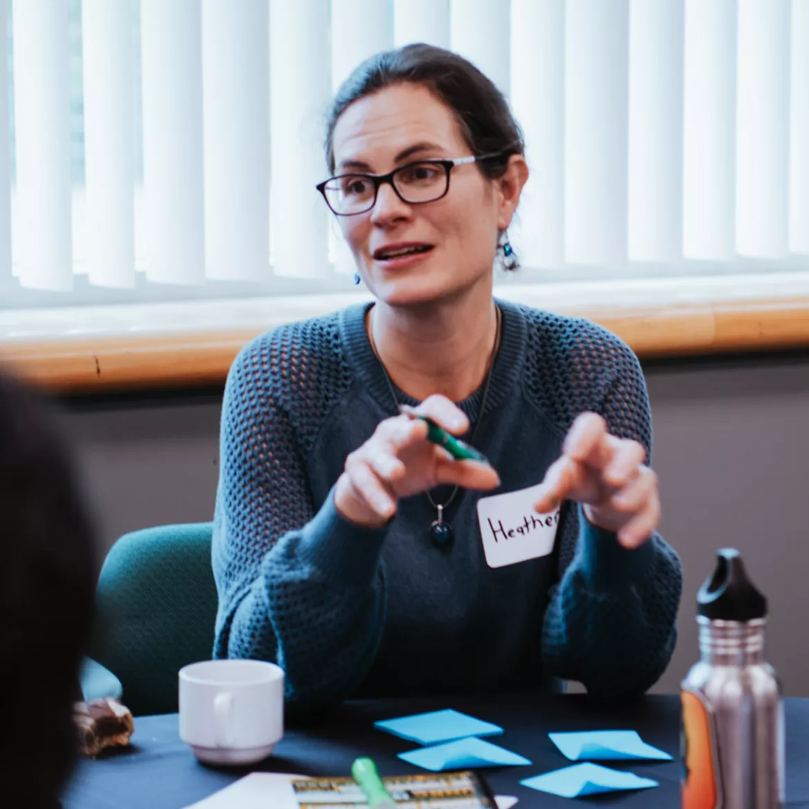 Person wearing green sweater gestures with hands while sitting at a table.