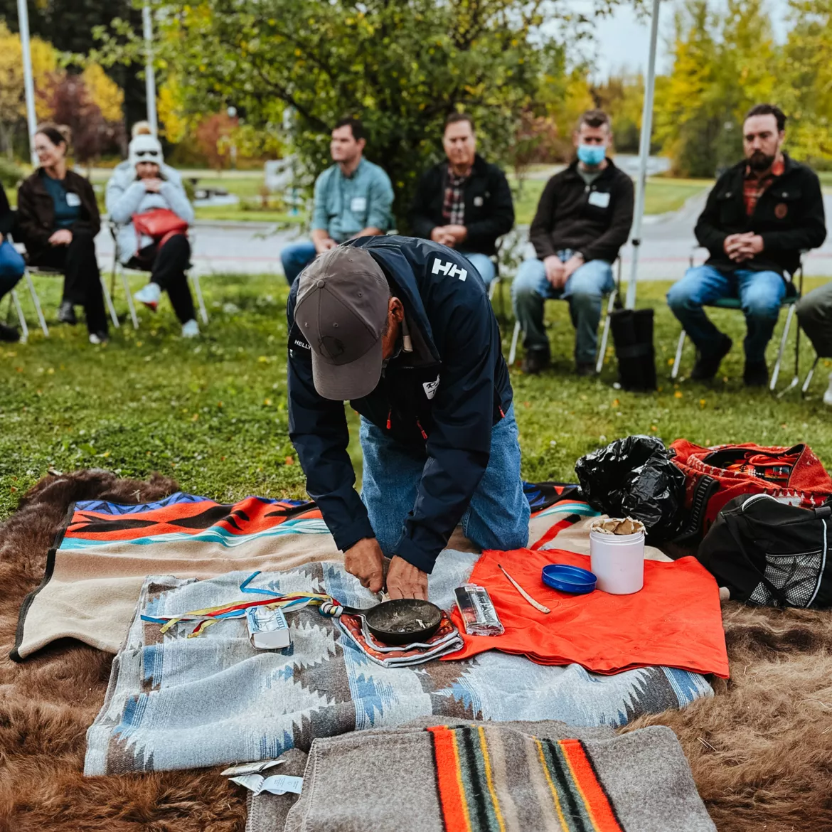 Person wearing hat, navy jacket and jeans kneels on a blanket preparing for a traditional opening ceremony.