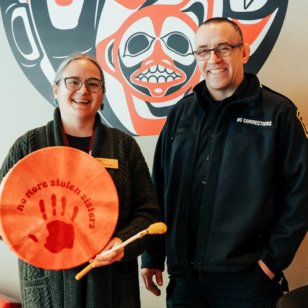 Two people stand together. Person on left holds hand drum.