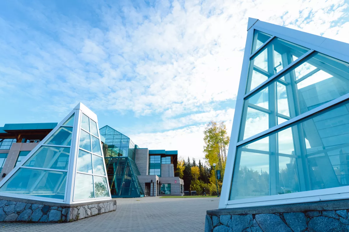 A view of the University of Northern British Columbia (UNBC) campus, featuring two large, pyramid-shaped glass structures in the foreground. These glass pyramids have white metal frames and rest on stone bases, standing prominently against a backdrop of the main campus building. The main building, visible in the background, also features modern architectural design with extensive use of glass and angular shapes. The sky is bright and partly cloudy, adding to the clean, contemporary feel of the scene. 