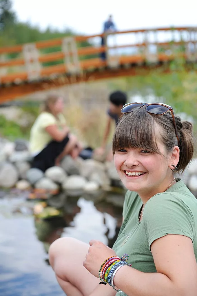 Student turned around and smiling at David Douglas Botanical Garden while students walk across bridge in background.