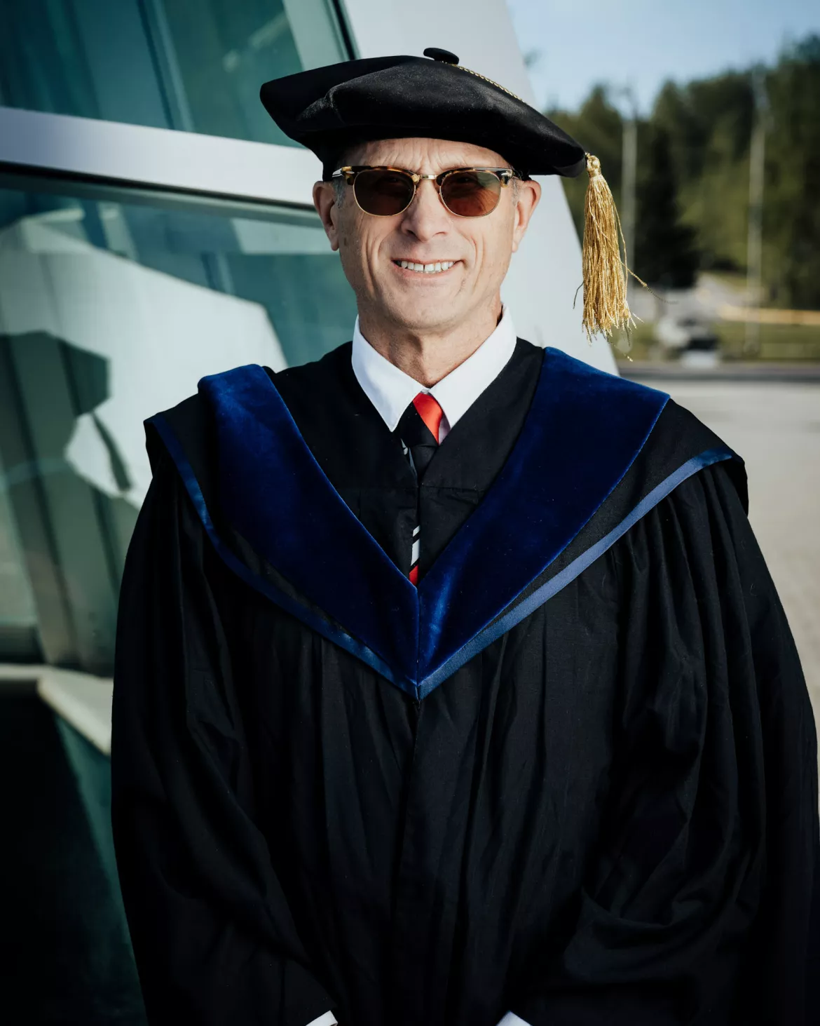 Person wearing dark glasses and black academic regalia and cap poses for photo outside.