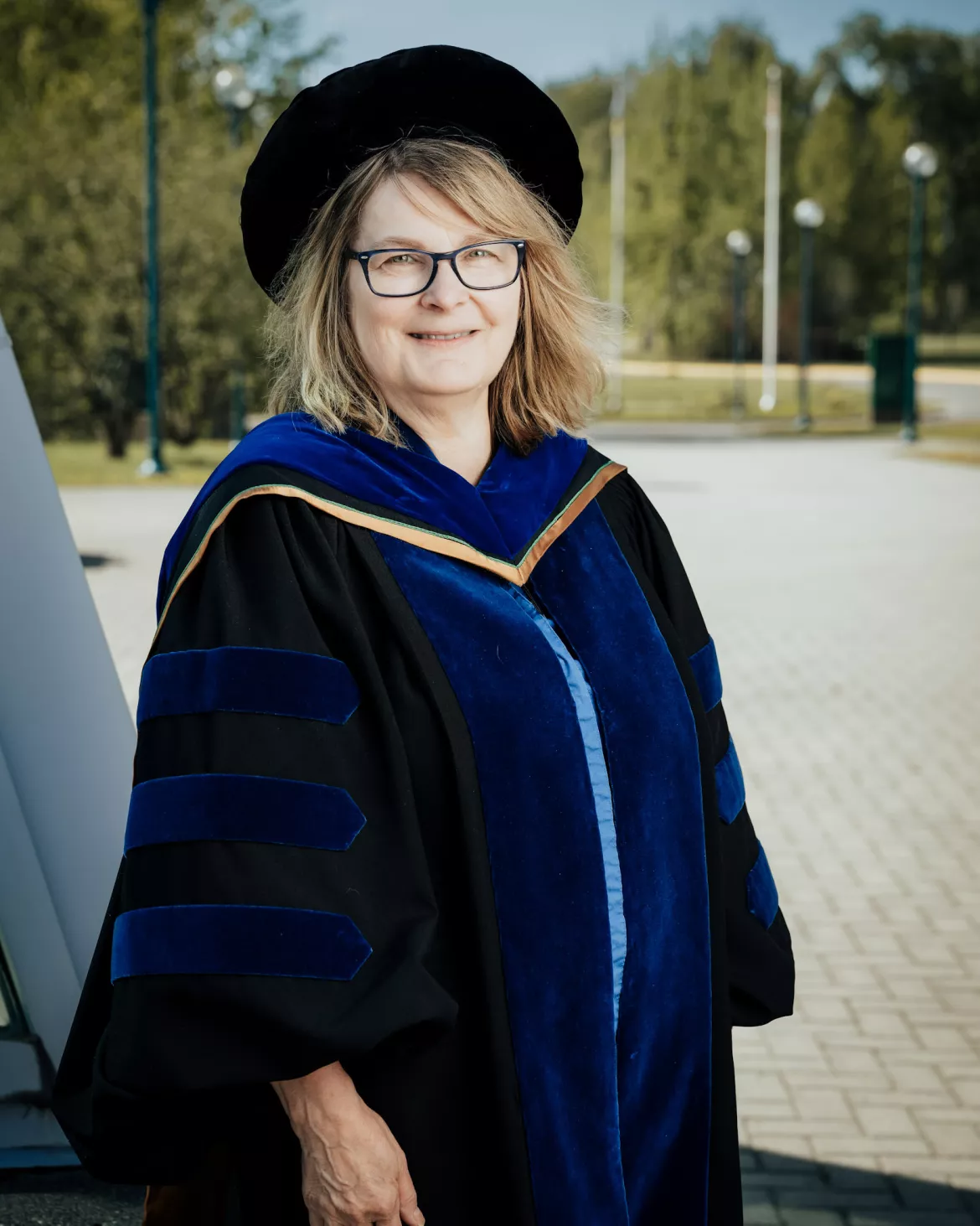 Person wearing black academic regalia with blue hood and trip poses outside.