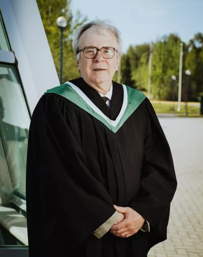 Person wearing black academic robe with white and green trim poses for photo outside.
