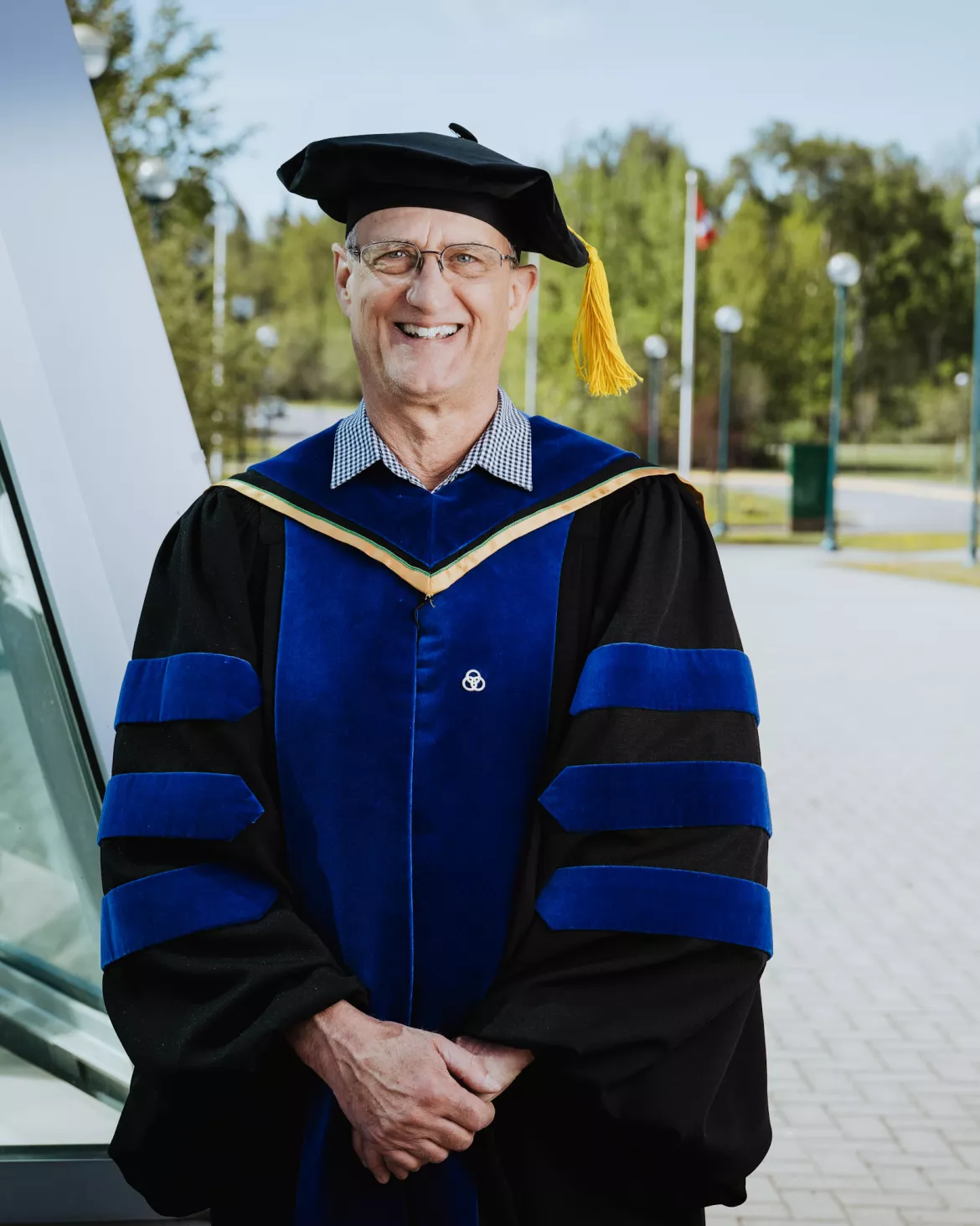 Person wearing black academic regalia, cap and blue trimmed hood poses for photo outside.