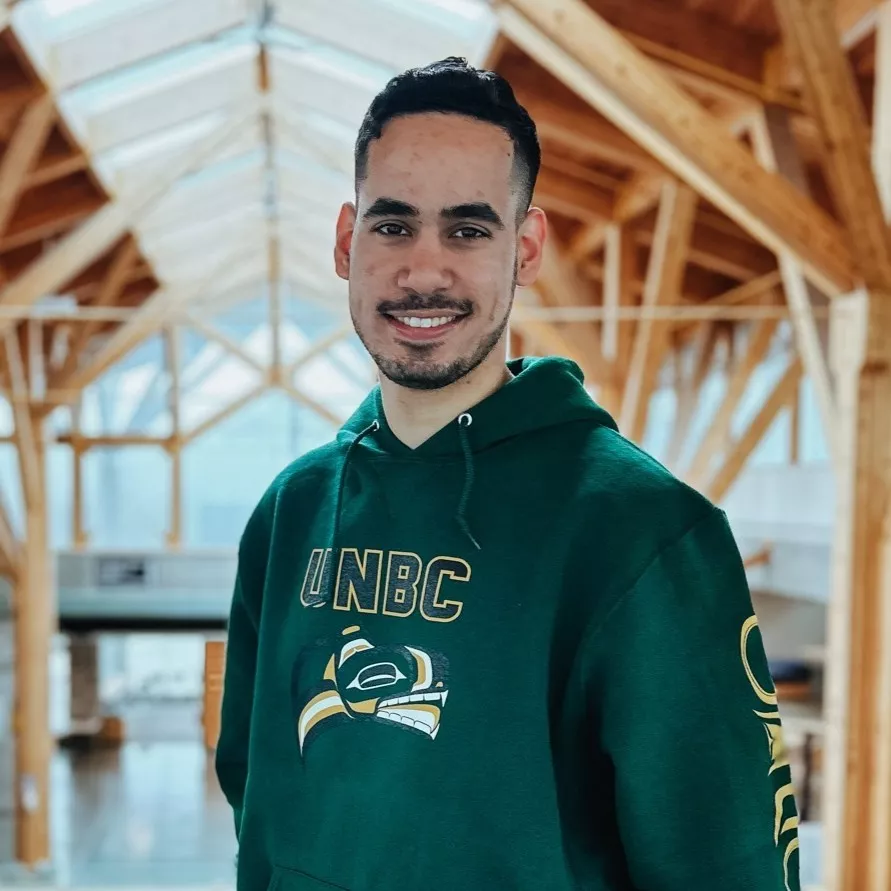 A student wearing a green UNBC hoodie with Indigenous-inspired artwork smiles in a bright, open atrium with wooden beams and large windows. The setting highlights the university’s distinctive architecture and welcoming environment.