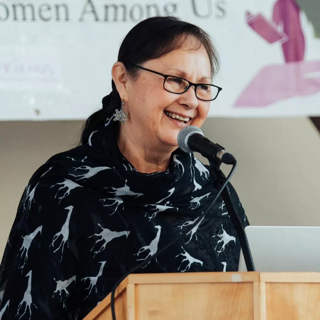 A woman wearing glasses and a patterned scarf smiles while speaking at a podium during an event. A banner in the background reads "Women Among Us," suggesting the occasion celebrates or honours women in the community.