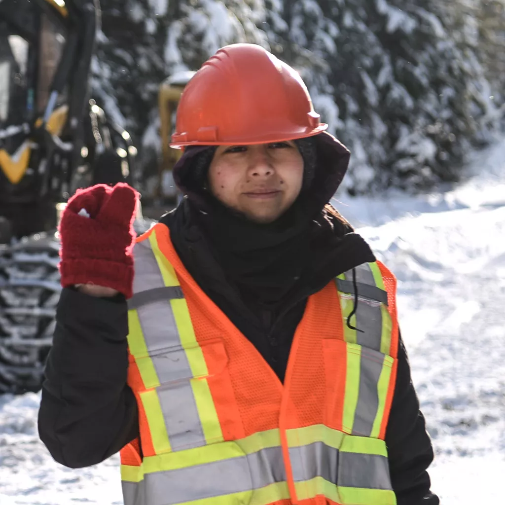 Person in orange safety vest and hard hat stands with red-mittened hand raised