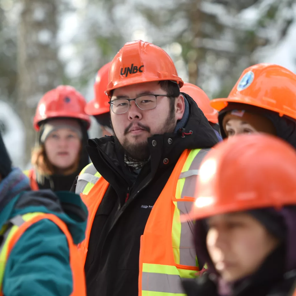 Group of students standing outdoors wearing orange hard hats and safety vests. Focus is on centre student.