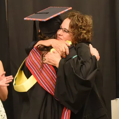 Two people hugging. Person on left wears black academic cap and gown with red Metis sash across their back. Person on right wears black.