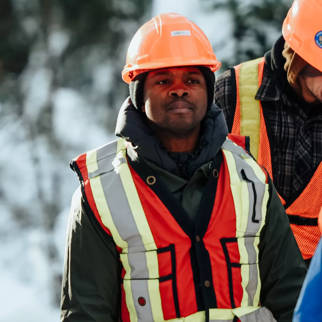 Person in orange safety vest and hard hat stands in snowy forest, another person stands on right side in background.