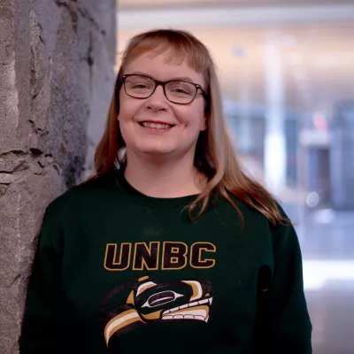 Student wearing a UNBC shirt with the Indigenous logo leans against a wall