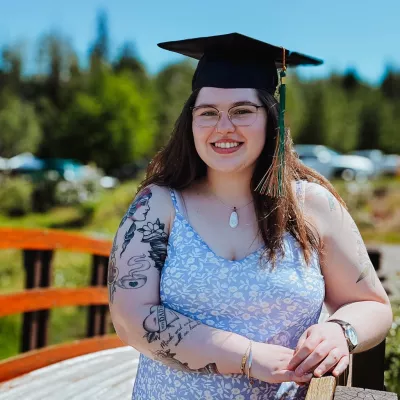 Student wearing floral sundress and black graduation cap with mortar board stands outside with a wooden bridge and greenery in the background.