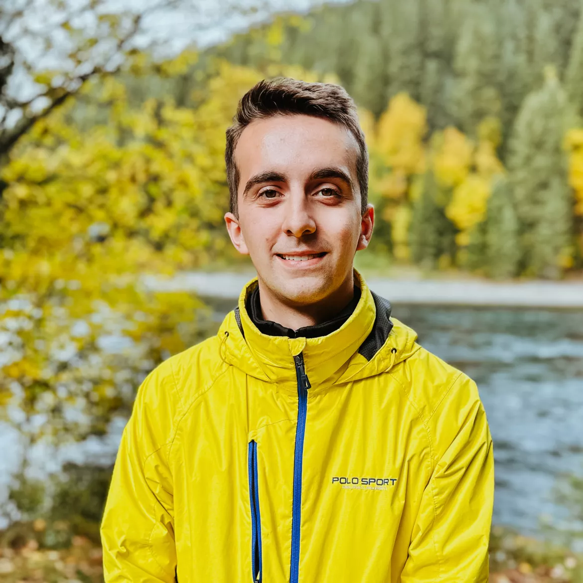 Person wearing yellow jacket stands on river shoreline with fall foliage in background.