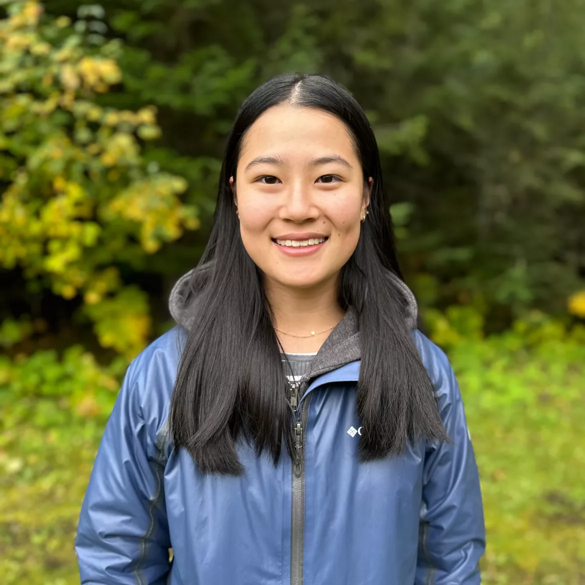 Person smiling, wearing a blue jacket outside in fall foliage.