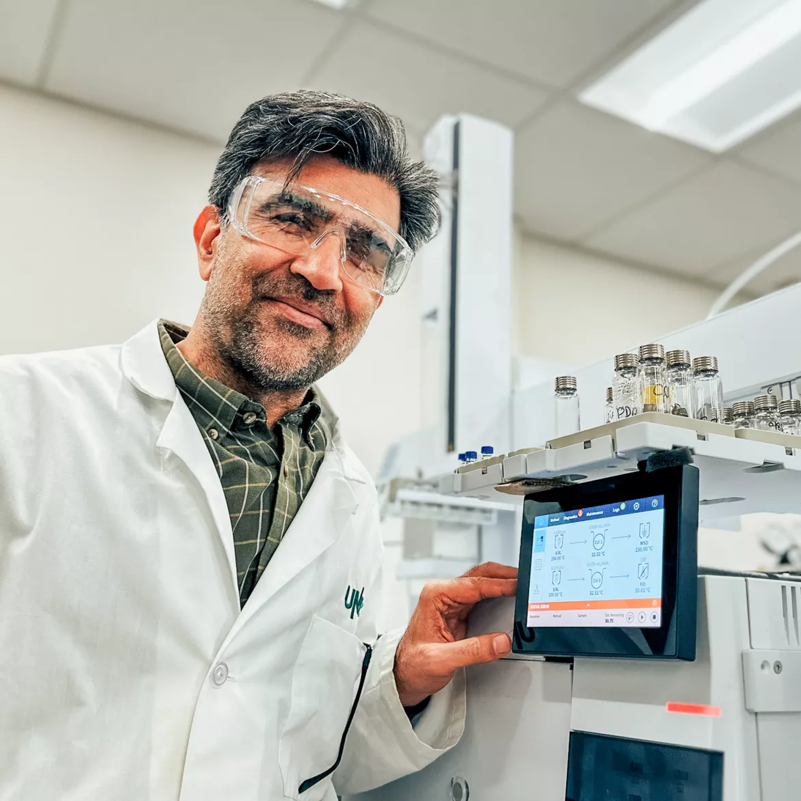 Person wearing plaid shirt and white lab coat leans on lab equipment with display panel.