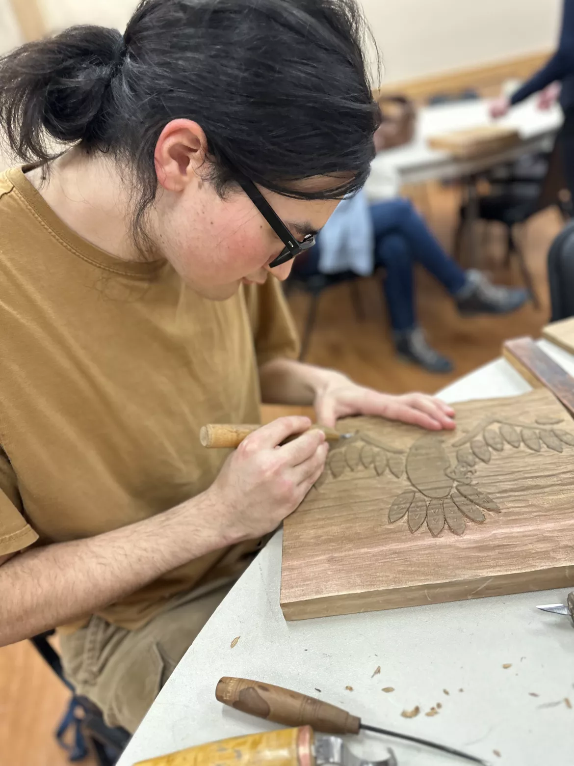 Man in tan t-shirt carves wooden plank at a table