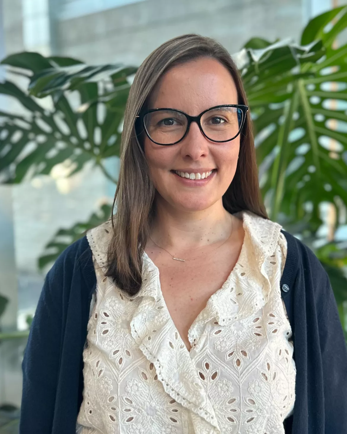 Person wearing glasses, white eyelet blouse and navy sweater stands in front of a plant.