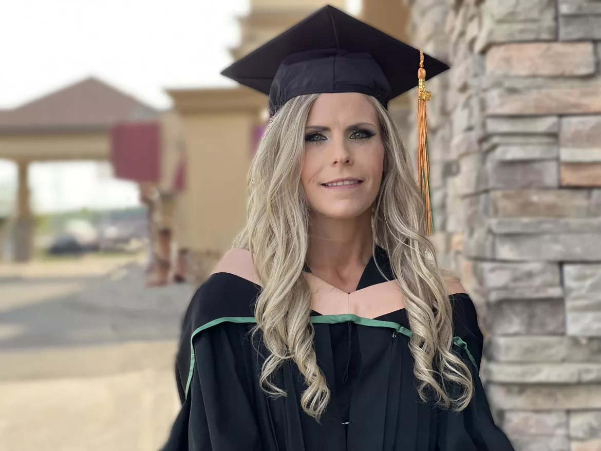 Student wearing black graduation gown with peach and green stripes and black cap with mortarboard stands outside next to a rock feature wall with buildings in background.