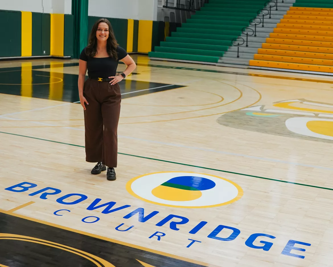 Person wearing black top and brown slacks stands in a gymnasium. Logo on flooring reads: Brownridge Court