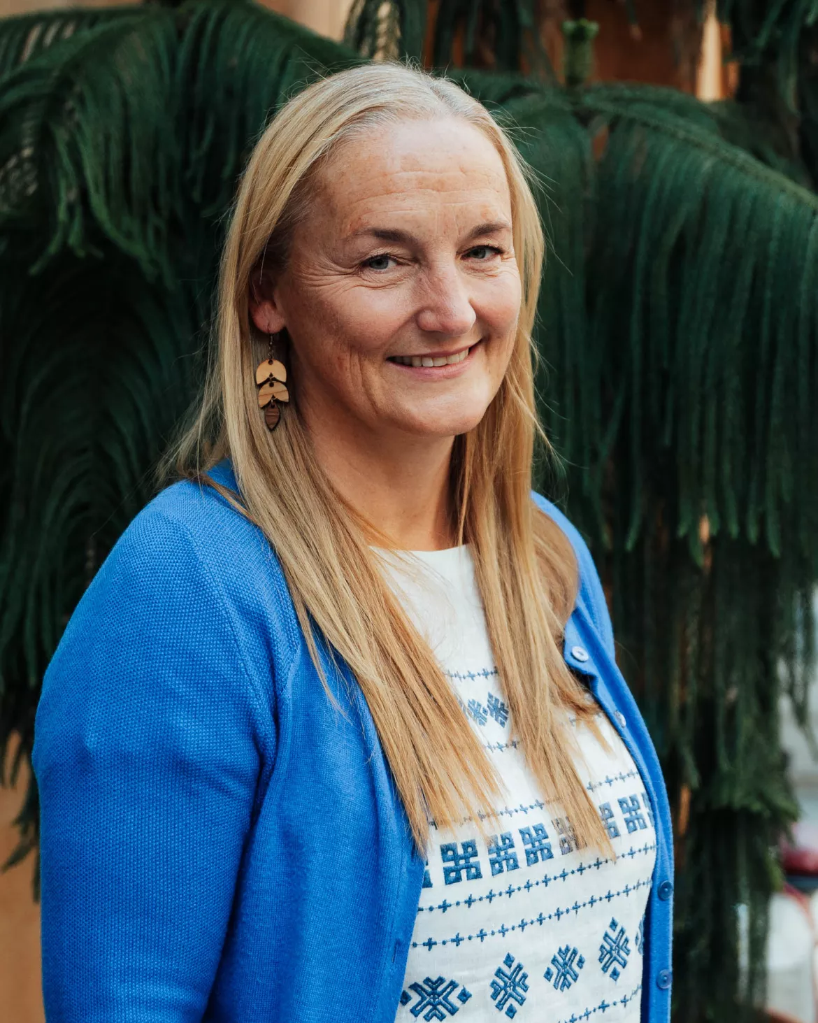 Dr. Caroline Sanders, wearing a blue cardigan and a white patterned shirt, smiles at the camera while standing outdoors in front of greenery.