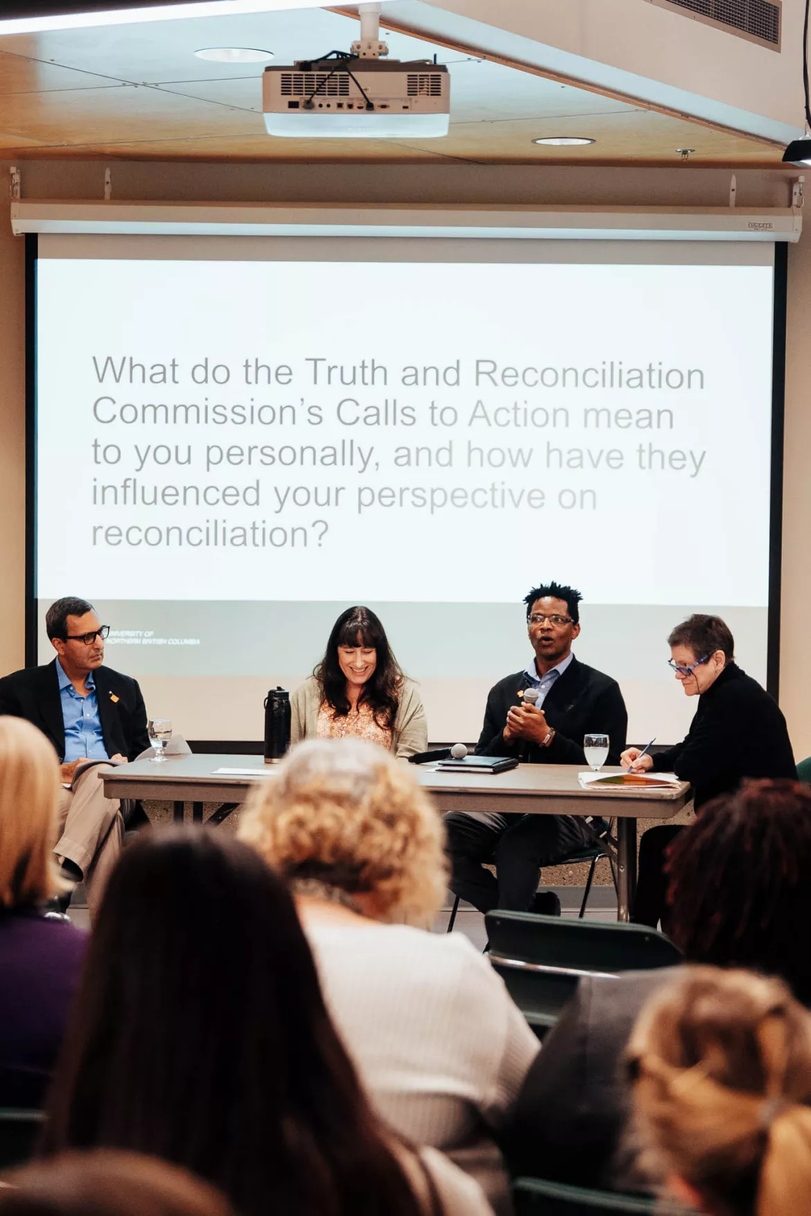 Four panelists sit at a table in front of a projected slide that reads, ‘What do the Truth and Reconciliation Commission’s Calls to Action mean to you personally, and how have they influenced your perspective on reconciliation?’ The panelists, from left to right, are Rahim Somani, Erica Hernandez-Read, Dr. Taru Manyanga, and Dr. Theresa Healy. They are engaged in discussion while an audience is visible in the foreground.