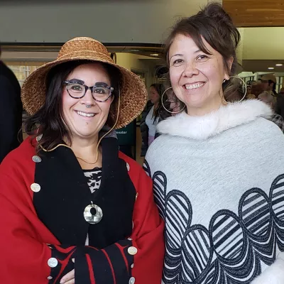 Person on left wears red button blanket and traditional woven hat. Person on right wears white and grey poncho.