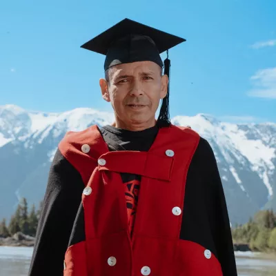Person wearing red and black button blanket and black mortarboard stands outside with mountains in background.